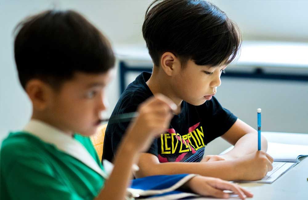Two young boys are sitting at a desk in a classroom writing on a piece of paper.