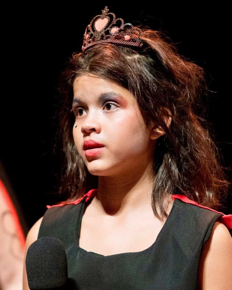 A young girl wearing a tiara and a black dress stands in front of a microphone