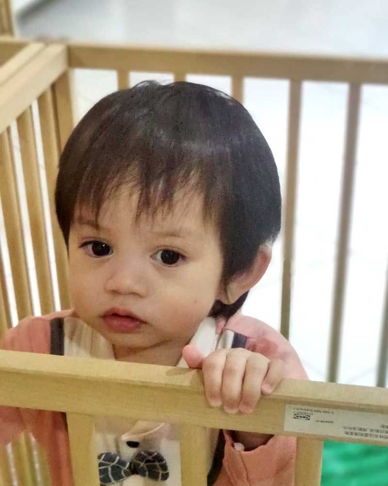 A baby is sitting in a wooden crib and looking at the camera.
