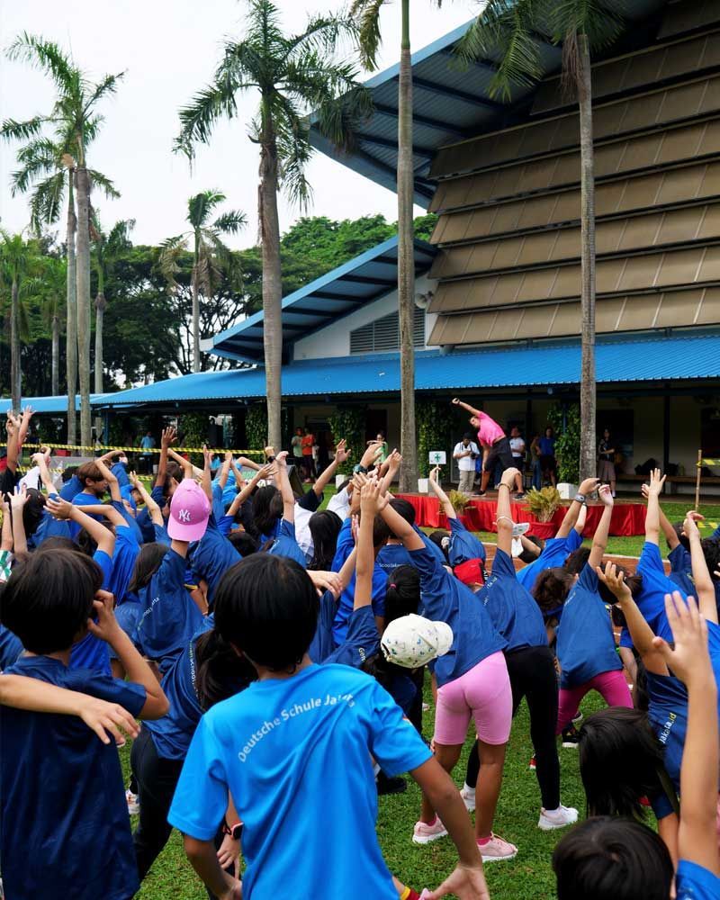 A group of children in blue shirts are stretching their arms in front of a building