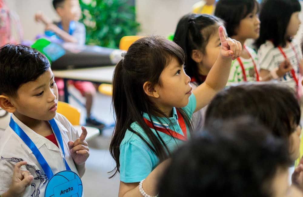 A group of children are sitting in a classroom raising their hands to answer a question.