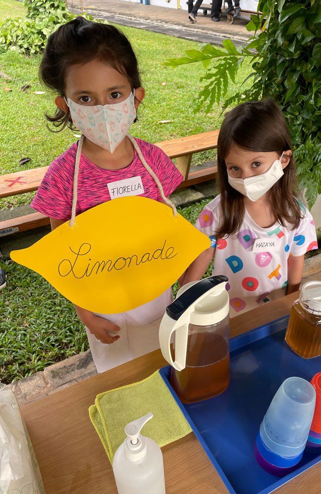 Two little girls wearing face masks are standing at a lemonade stand.