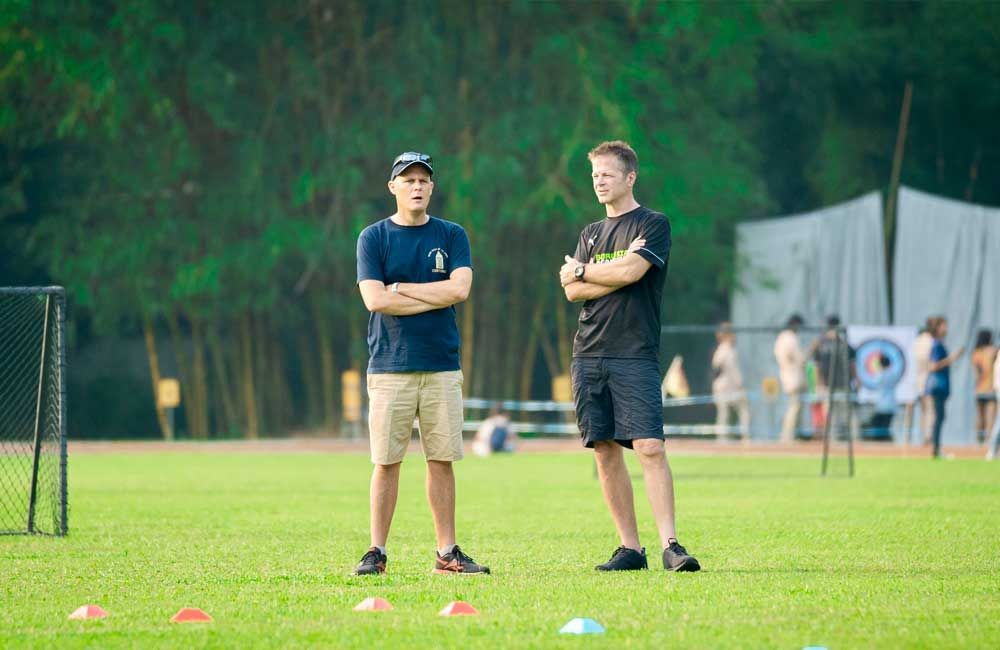 Two men are standing on a soccer field with their arms crossed.