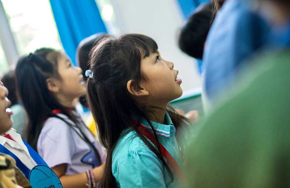 A group of children are sitting in a classroom looking up.