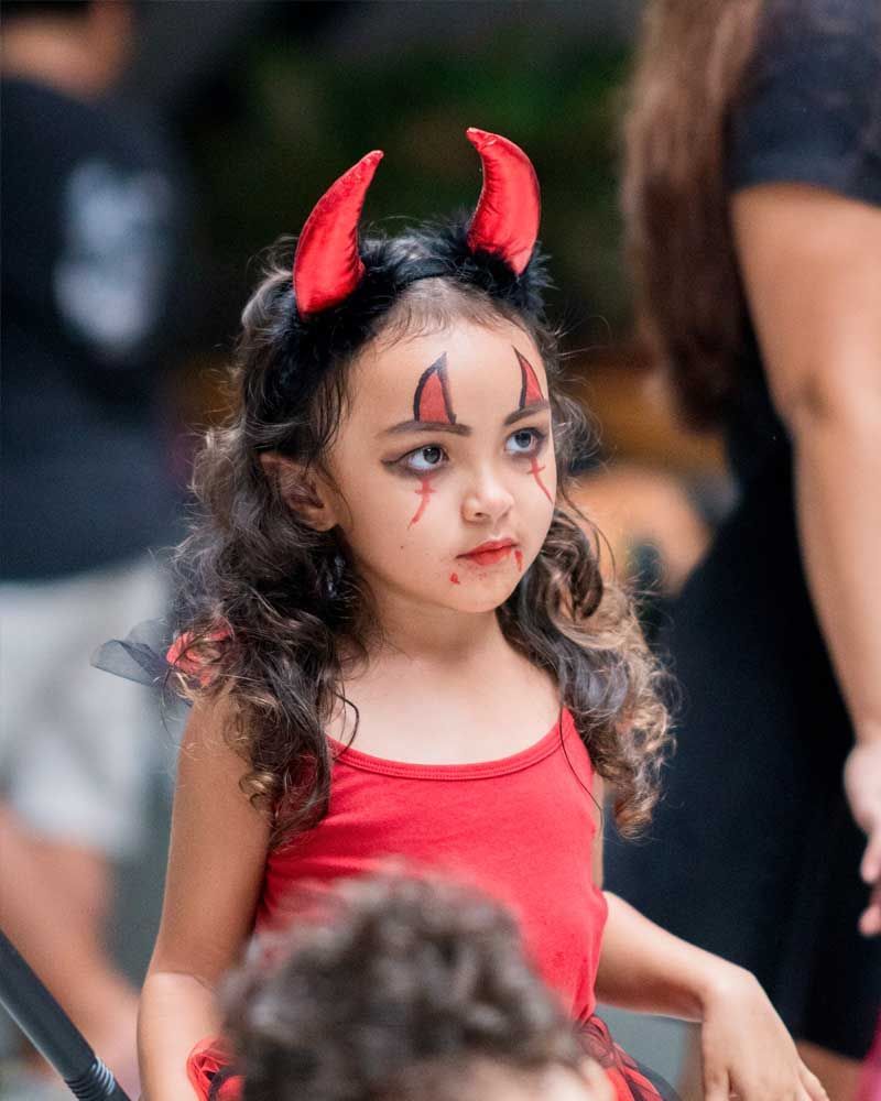 A young girl in a red devil costume with horns and face paint celebrates Halloween