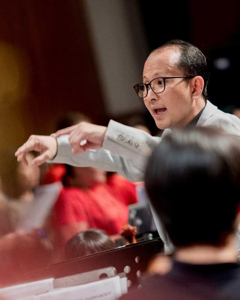 A man in a suit and glasses is standing in front of a choir.