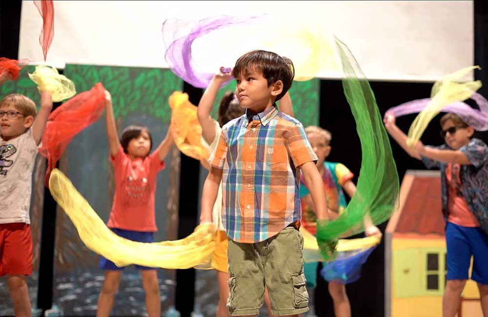 A group of children are dancing on a stage with colorful scarves.
