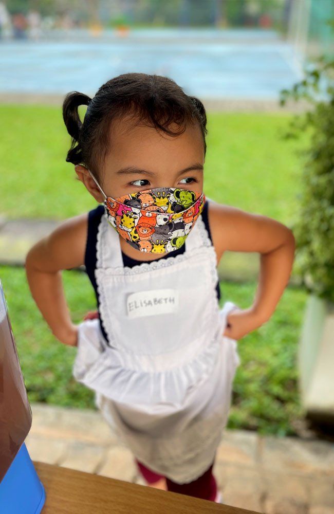 A little girl wearing a mask and an apron is standing with her hands on her hips.