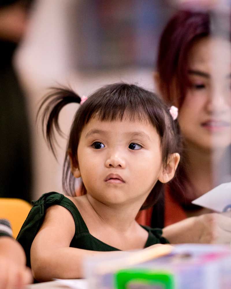 A little girl is sitting at a table with a woman behind her.