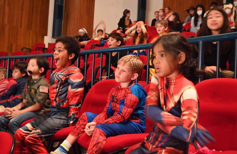 A group of children in superhero costumes are sitting in red chairs.