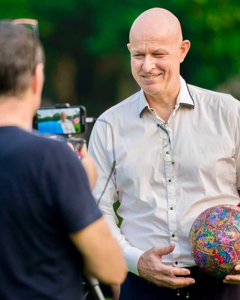A man in a white shirt is holding a soccer ball and smiling while another man takes a picture of him.