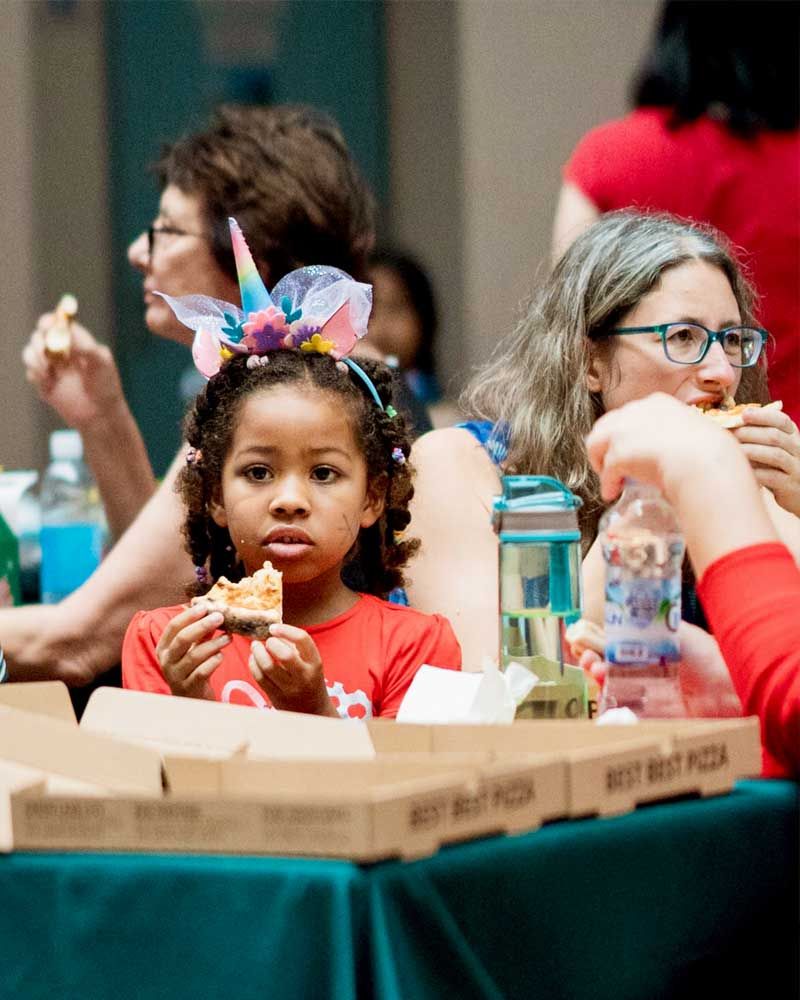 A little girl wearing a unicorn headband is sitting at a table eating pizza.