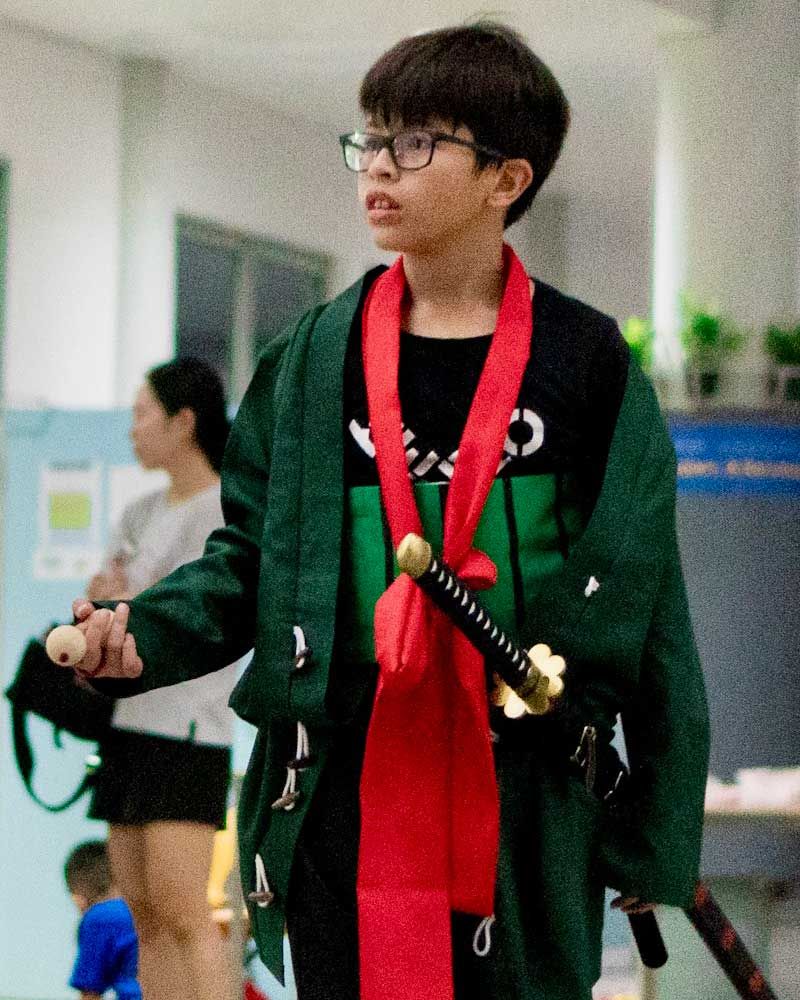 A boy dressed as a samurai with a green jacket, red scarf, and wooden sword takes part in a Halloween