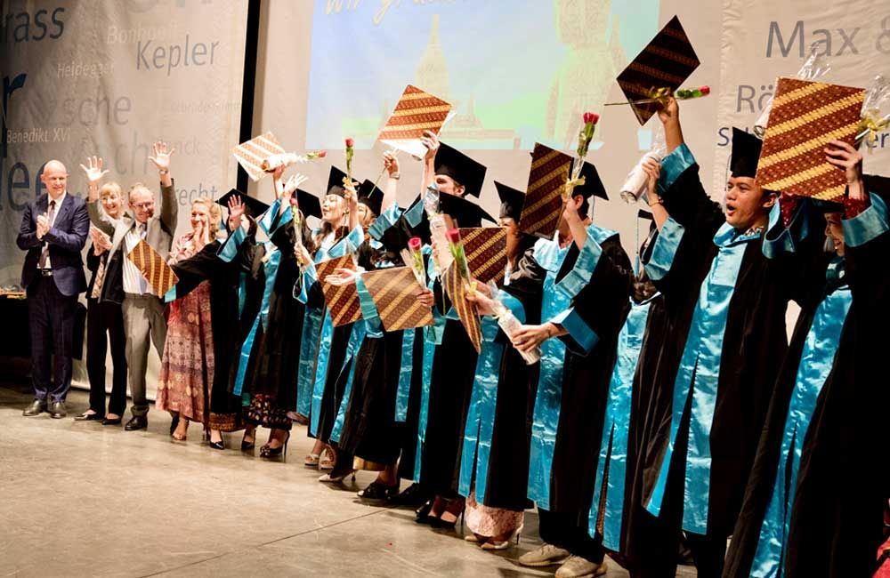 A group of graduates are standing on a stage holding up their caps and gowns.
