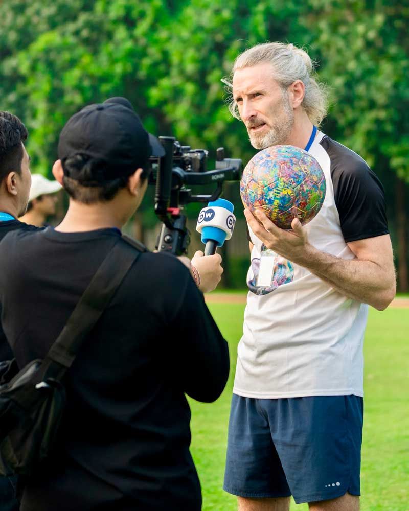 A man is holding a soccer ball and talking to a cameraman.