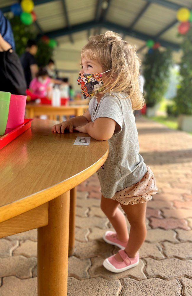 A little girl wearing a face mask is standing next to a table.