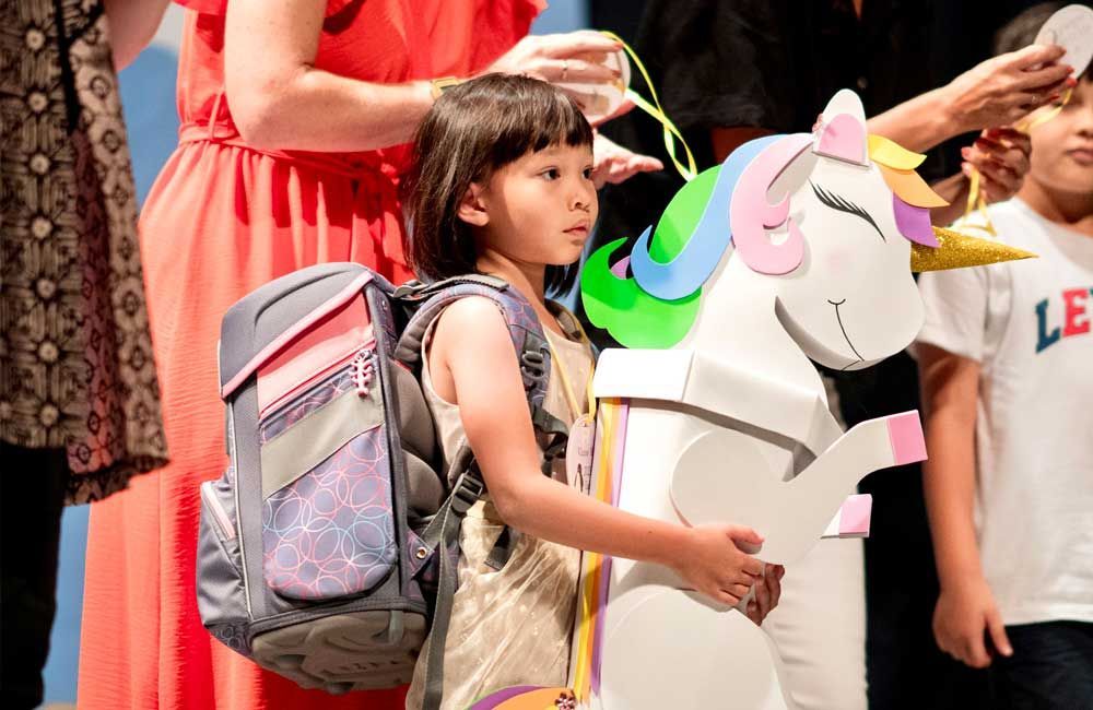 A little girl with a backpack is standing next to a cardboard unicorn.