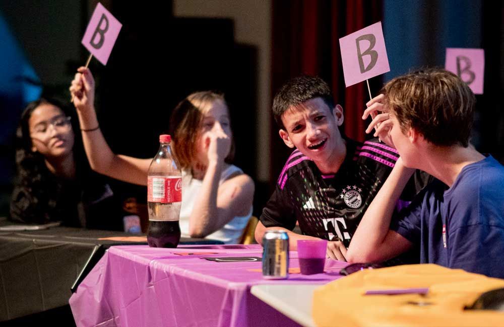 A group of young people are sitting at a table holding up signs with the letter b on them.