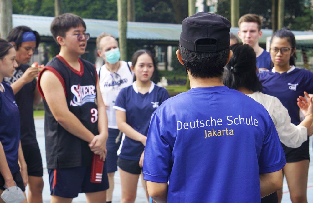 A group of people are standing around a man wearing a blue shirt that says deutsche schule jakarta