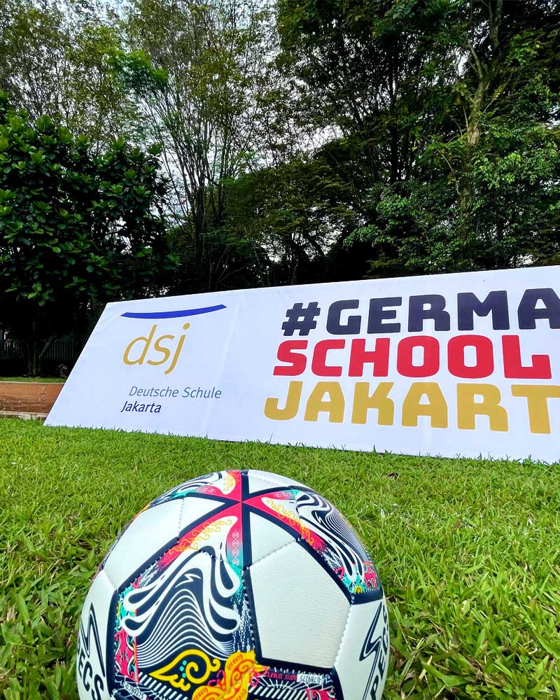 A soccer ball is sitting in the grass in front of a sign that says germa school jakarta