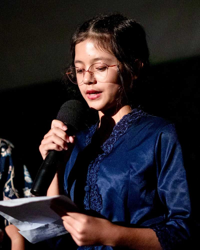 A young girl wearing glasses is holding a microphone and reading a piece of paper