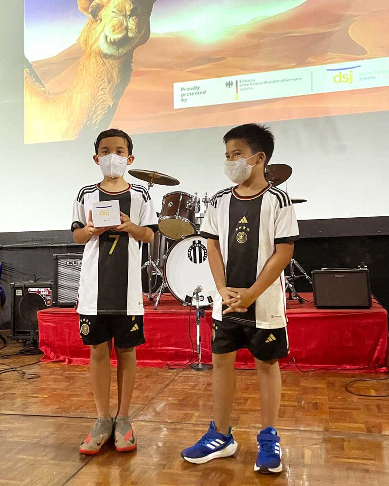 Two young boys wearing face masks are standing on a stage in front of a drum set.