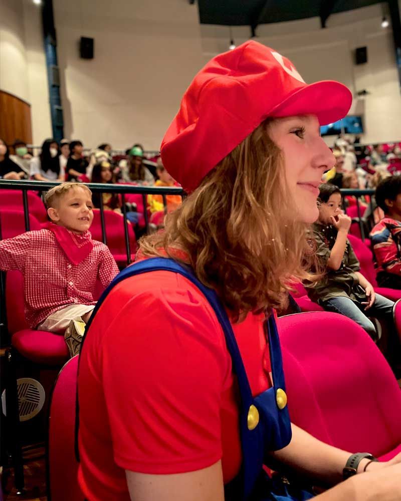 A woman in a red hat and overalls is sitting in a crowded auditorium.