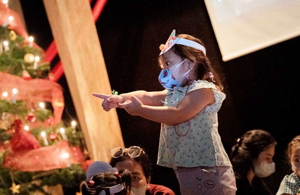 A little girl wearing a mask is pointing at something in front of a christmas tree.