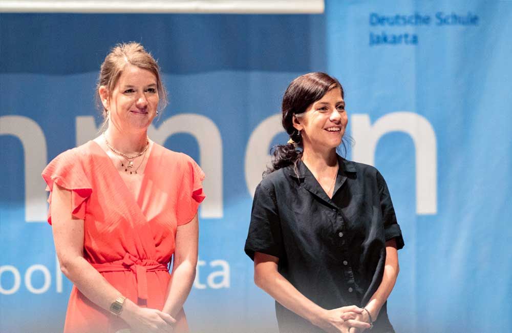 Two women are standing next to each other in front of a sign that says deutsche schule jakarta