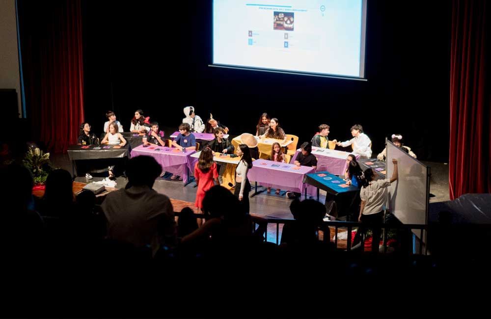 A group of people are sitting at tables on a stage in front of a large screen.