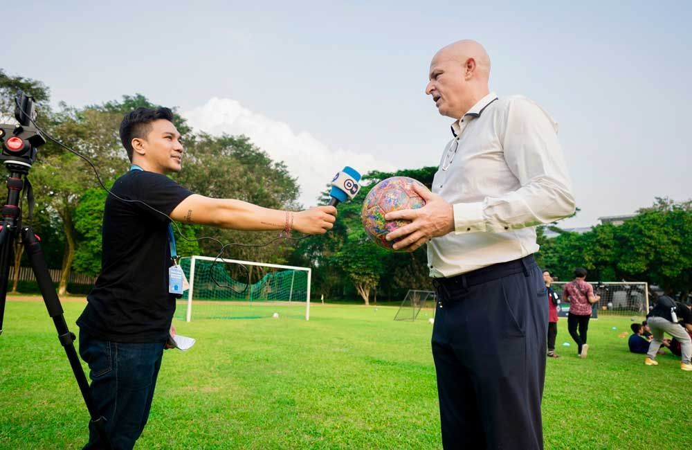A man is holding a soccer ball and talking to another man with a microphone.