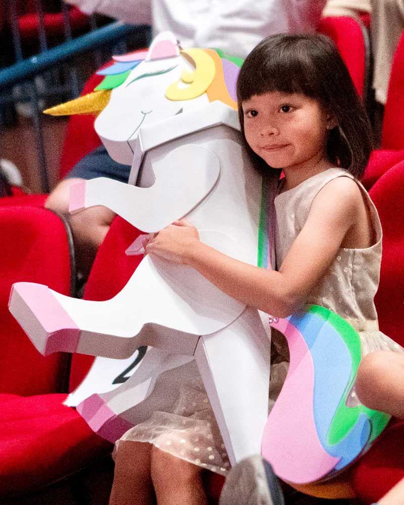 A little girl hugging a large unicorn cutout while sitting in a red auditorium seat.