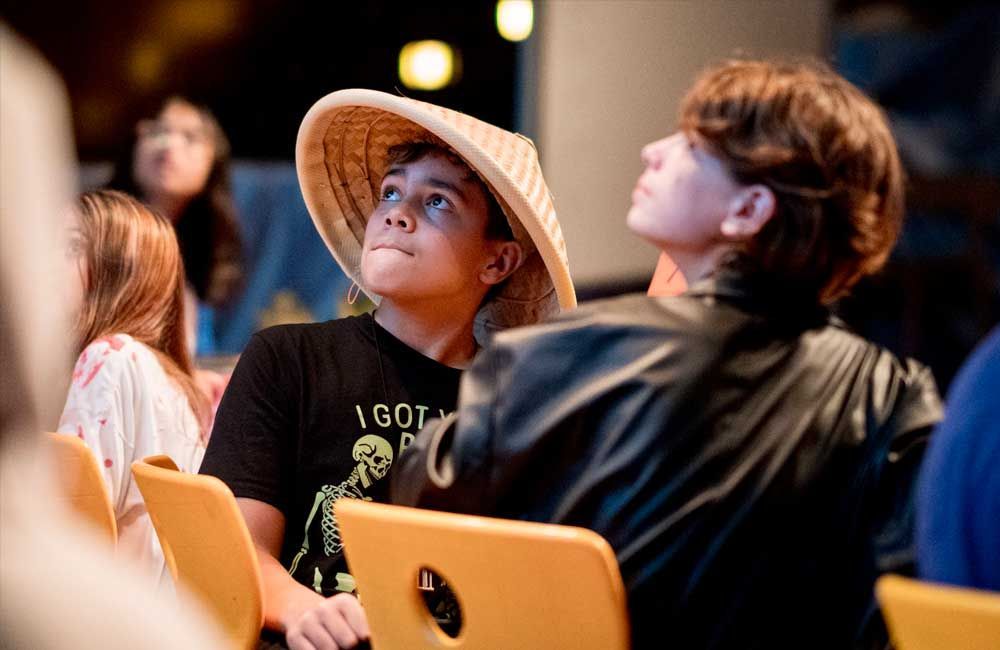 A boy wearing a conical hat is sitting in a chair looking up.