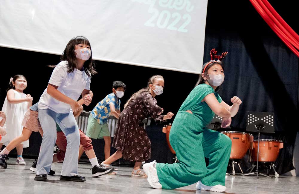 A group of children wearing face masks are dancing on a stage.