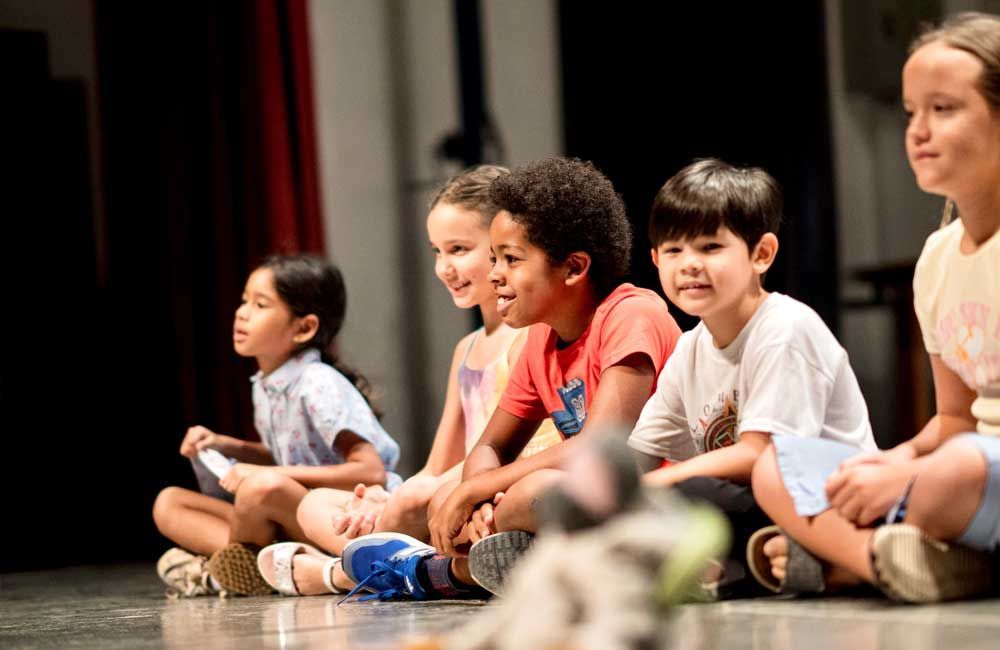 A group of children are sitting on the floor on a stage.