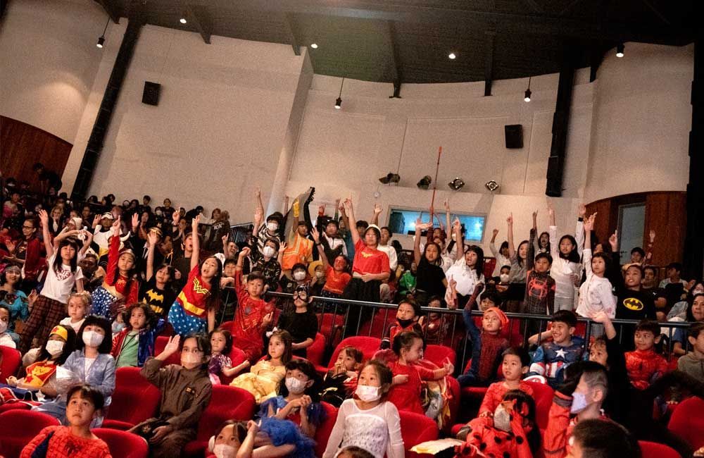 A large group of people are sitting in a theater with their hands in the air.