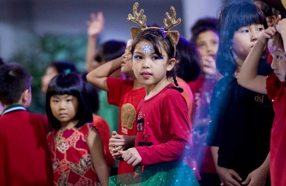 A little girl wearing a reindeer headband is standing in a crowd of children.