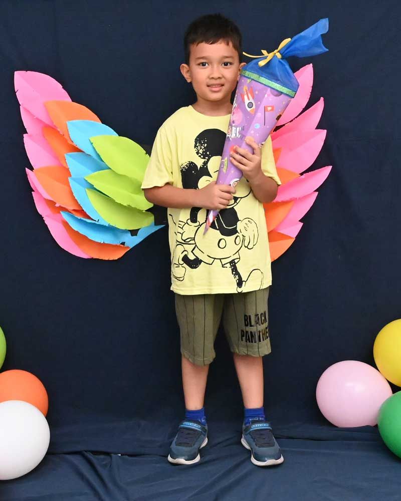 A young boy holding a decorated torch, standing in front of colorful wings at a school event