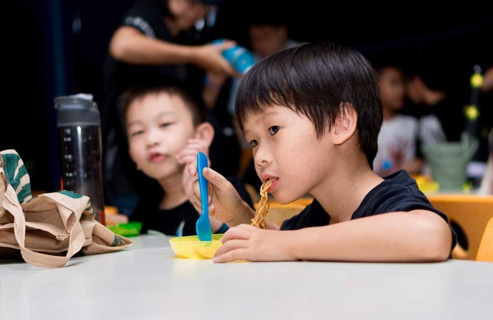 A young boy is sitting at a table eating noodles with a spoon.