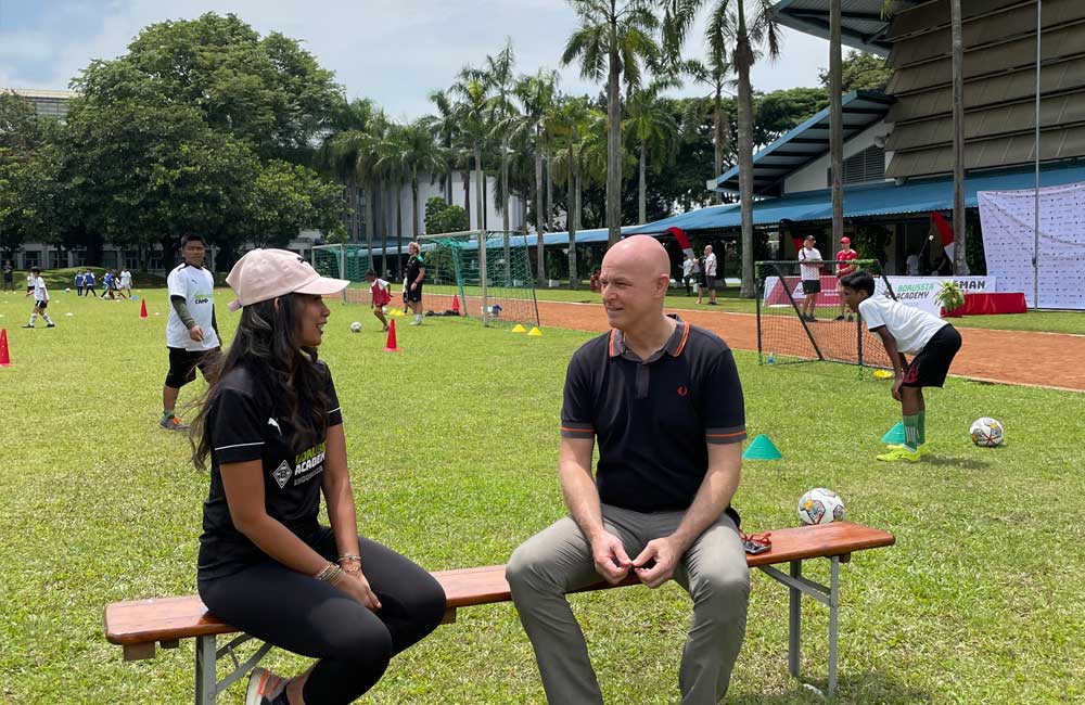 A man and a woman are sitting on a bench in a field talking to each other.