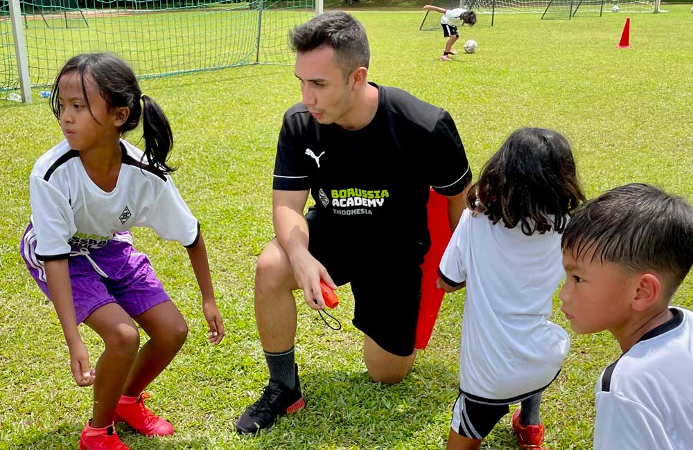 A man is kneeling down next to a group of children on a soccer field.