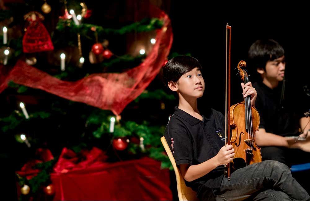 A boy is playing a violin in front of a christmas tree.