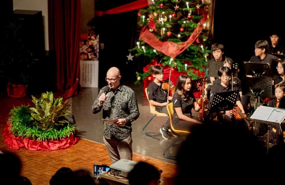 A man is standing on a stage with a microphone in front of a christmas tree.