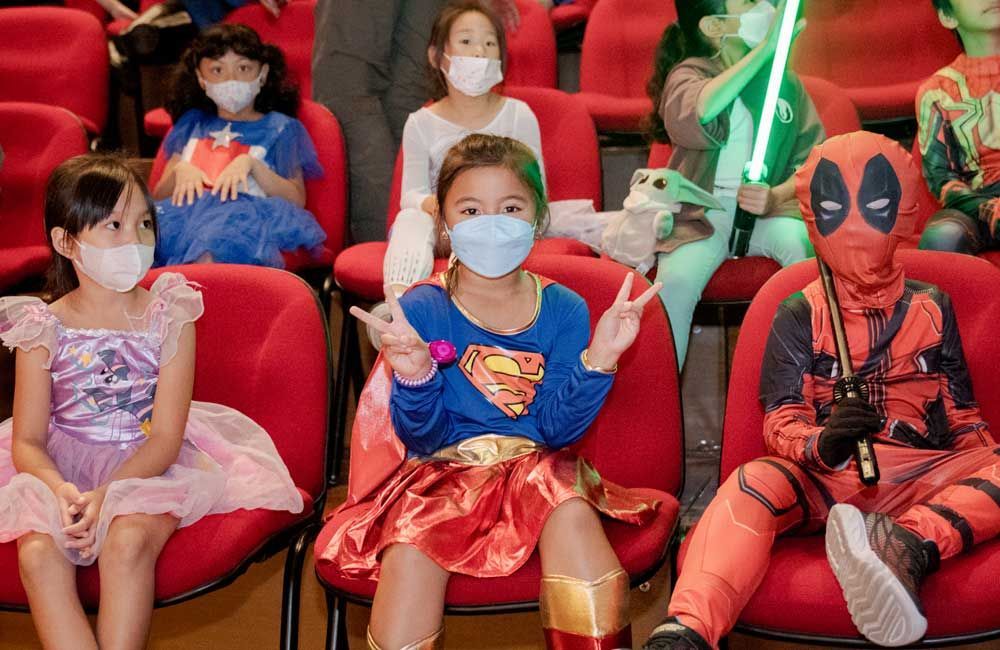 A group of children dressed in costumes are sitting in red chairs.