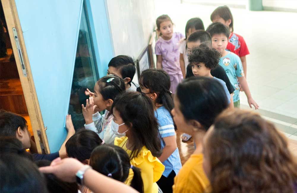 A group of children are standing in a line looking through a glass door.