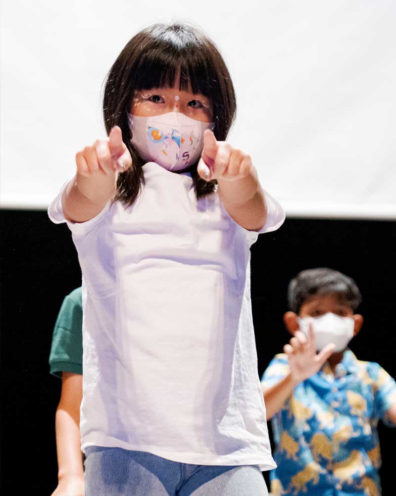 A little girl wearing a face mask is pointing at the camera.