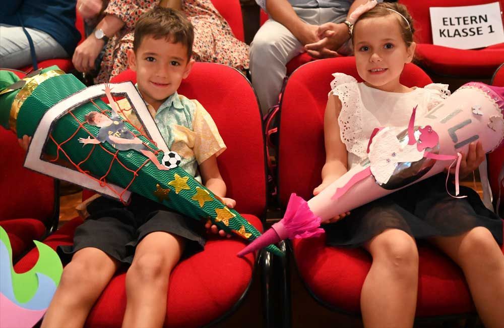 A boy and a girl are sitting in red chairs holding cones and a sign that says eltern klasse 1
