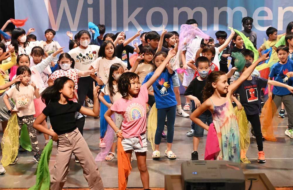 A group of children are dancing on a stage in front of a sign that says willkommen.
