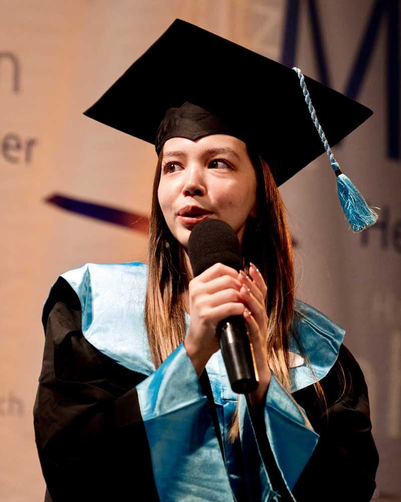 A woman in a graduation cap and gown is speaking into a microphone