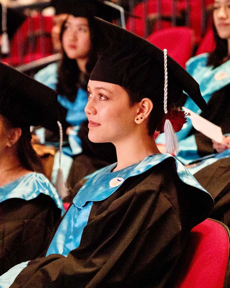 A woman in a graduation cap and gown sits in a row of people